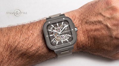 Michel Herbelin skeleton watch in a dark grey cushion case with an exposed movement, luminous baton hands and a grey mesh bracelet on the wrist