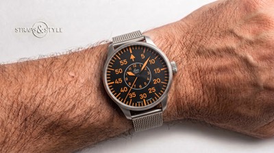 Laco pilot watch with a black dial, orange numerals and hands, and a steel mesh bracelet on the wrist