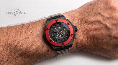 Hublot skeleton chronograph with a matte black case, red bezel and yellow date window on a Straps&Style mesh bracelet