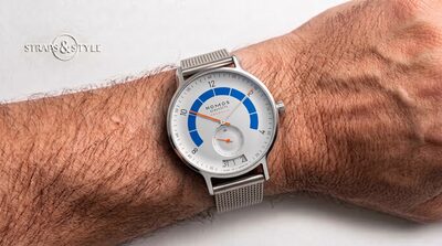 NOMOS Glashütte Autobahn with a silver-white dial, a bright blue speedometer-style arc, neon-orange hands and a curved date window at six on a Straps&Style mesh bracelet